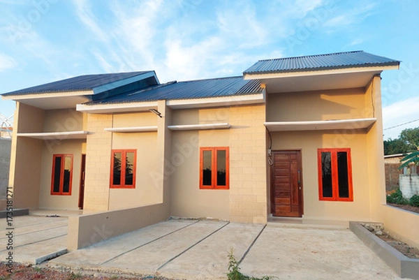 Fototapeta A simple permanent house with a modern minimalist design, beige walls, wooden doors, and red-framed windows, viewed from the front under a clear blue sky.