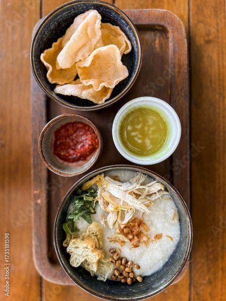 Fototapeta Traditional Indonesian chicken porridge or Bubur Ayam served with crackers, chili sauce, green sauce, and toppings on a wooden tray over rustic wooden table.