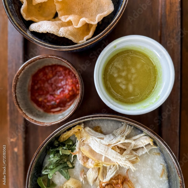 Fototapeta Traditional Indonesian chicken porridge or Bubur Ayam served with crackers, chili sauce, green sauce, and toppings on a wooden tray over rustic wooden table.
