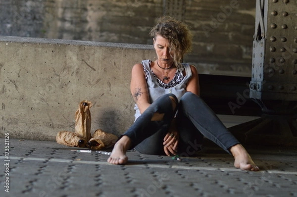 Fototapeta a hopeless punk woman under a bridge with bottles of alcohol and other drugs around her 