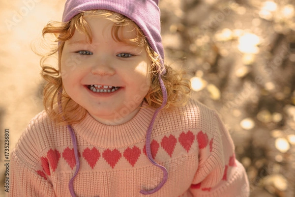 Fototapeta A cheerful toddler with curly hair and a purple hat smiles brightly outside, surrounded by a warm, sunny atmosphere and golden leaves in the background.