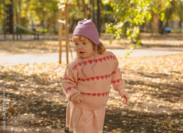 Fototapeta A young child with curly hair runs joyfully through a park covered in colorful autumn leaves. She wears a cozy sweater and a cute hat, enjoying the beautiful sunny day.