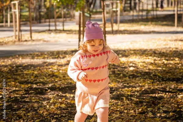 Fototapeta A young child with curly hair runs joyfully through a park covered in colorful autumn leaves. She wears a cozy sweater and a cute hat, enjoying the beautiful sunny day.