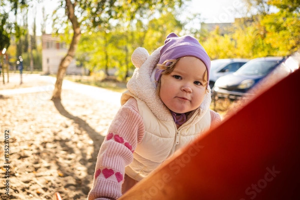 Fototapeta A happy toddler in a pink sweater slides down a playground slide, laughing with joy. The setting features green trees and colorful play equipment, showing a typical day at the park.