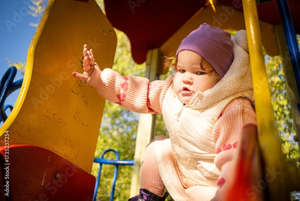 Fototapeta A young child enjoys playing on a playground in a lovely park. Dressed warmly, the child smiles while exploring the climbing structure under a clear blue sky.