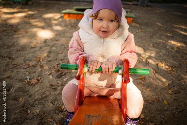 Fototapeta Child plays on a red playground toy in a sunny park surrounded by trees and benches