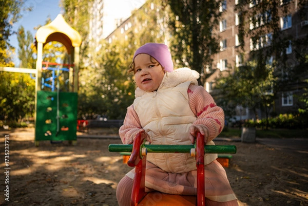 Fototapeta Child plays on a red playground toy in a sunny park surrounded by trees and benches
