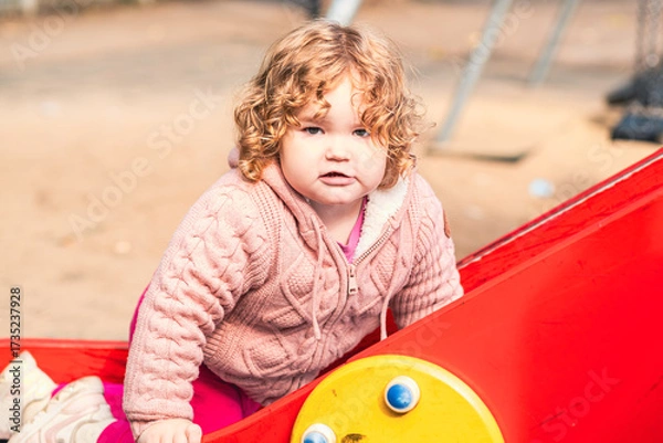Fototapeta A happy toddler in a pink sweater slides down a playground slide, laughing with joy. The setting features green trees and colorful play equipment, showing a typical day at the park.