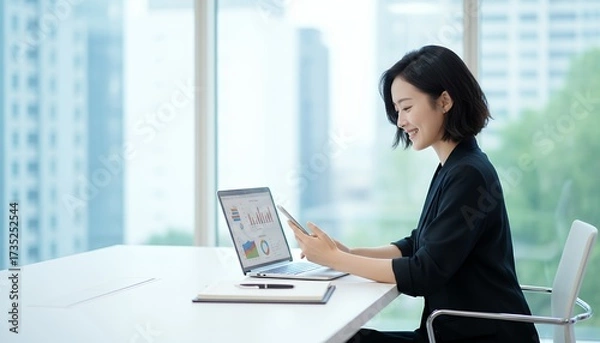 Fototapeta PCとスマホを手にオフィスで微笑む女性