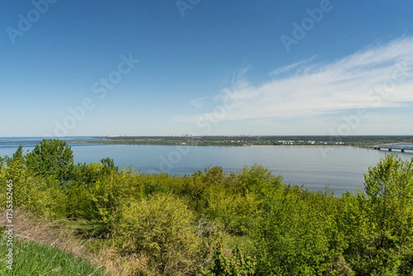 Fototapeta Wide Volga River under a clear blue sky, framed by green trees. City buildings and a bridge are visible on the horizon.