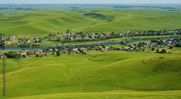 Fototapeta Expansive green landscape with rolling hills and a river bisecting a small settlement under a clear sky