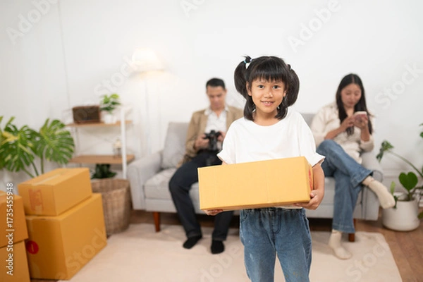 Fototapeta New Beginnings: A smiling young girl in casual attire carries a cardboard box, symbolizing moving to the new home, standing in the cozy living room where parents are sitting on sofa.