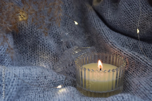 Fototapeta A lit candle on warm blanket decorated with garland. Cozy atmosphere with a candle. 