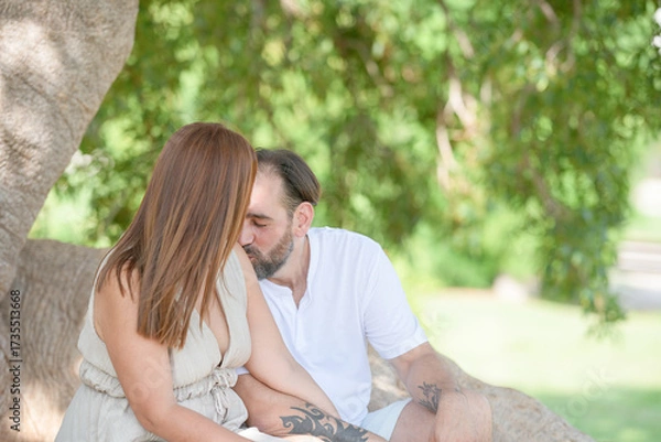 Fototapeta A man and a woman are sitting on a tree branch, kissing each other. Scene is romantic and intimate