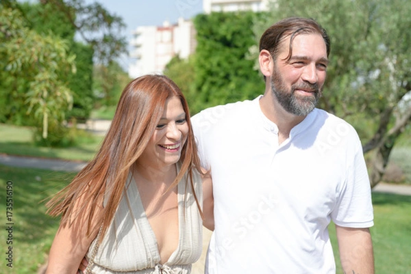 Fototapeta A man and a woman are walking together in a park. The man is wearing a white shirt and the woman is wearing a white dress. They are smiling and seem to be enjoying each other's company