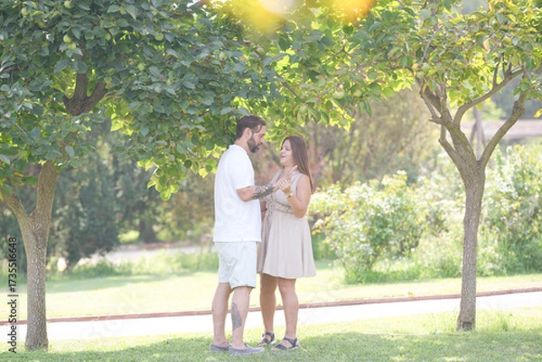 Fototapeta A couple is standing in a park, with trees in the background. The man is wearing a white shirt and shorts, while the woman is wearing a dress