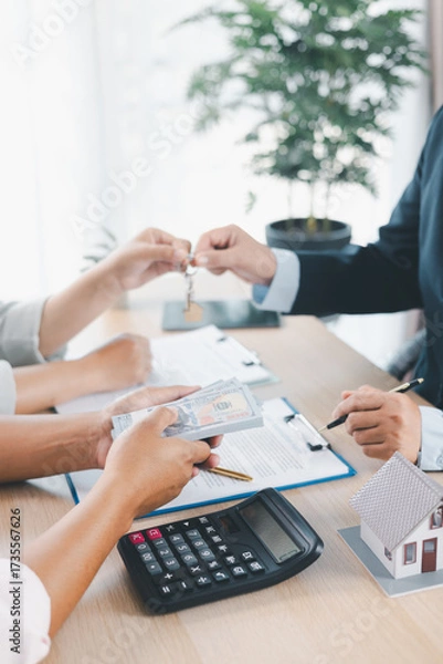 Fototapeta Close up of real estate agent handing house key to client with contract paperwork, calculator and house models on desk, concept of home ownership, mortgage loan, property investment and success.
