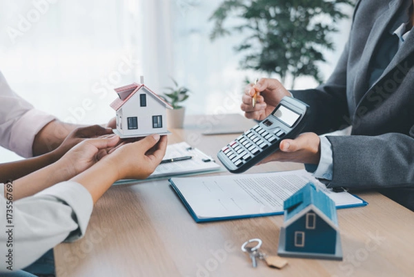 Fototapeta Close up of real estate agent handing house model to client with calculator and contract paperwork on desk, concept of mortgage loan, home ownership, property investment and housing agreement.