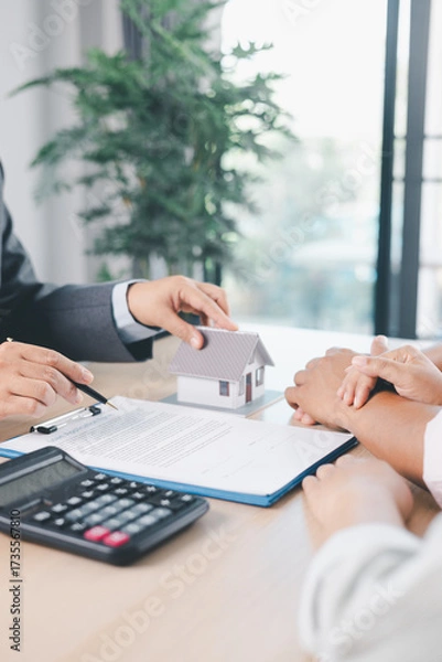 Fototapeta Close up of real estate agent showing small house model to client with calculator, contract paperwork on desk, concept of mortgage loan, home ownership, property investment agreement. mortgage loan
