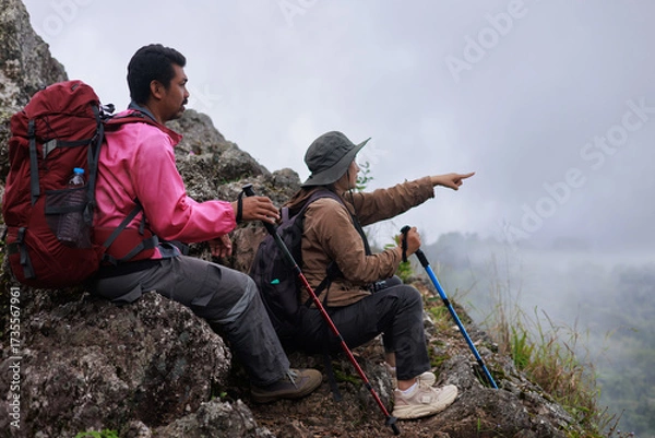 Fototapeta Two hikers with backpacks and trekking poles resting on rocky mountain, woman pointing forward, symbol of teamwork, adventure, exploration, outdoor lifestyle and travel inspiration in nature.