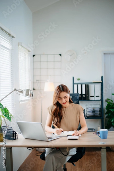 Fototapeta Asian businesswoman multitasking with phone and notebook in bright office. Perfect for business, communication, consulting, productivity, and professional work themes.