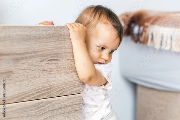Fototapeta Baby holding onto wooden furniture while standing indoors, looking thoughtful and curious, symbol of growth, exploration and early childhood development