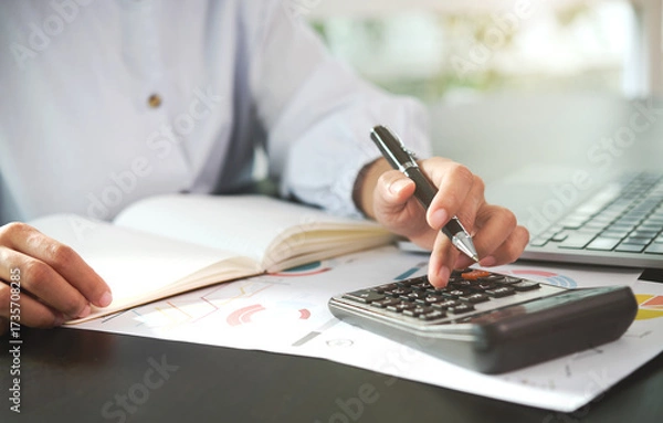 Fototapeta Businessperson using calculator with financial charts and laptop on desk office