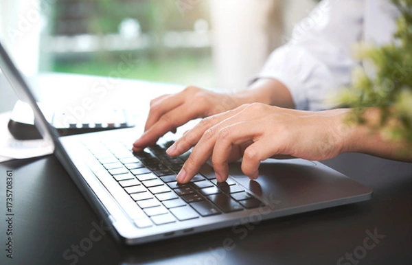 Fototapeta Close-up of hands typing on laptop keyboard with blurred green plant on table background