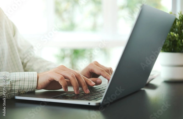 Fototapeta Close-up of hands typing on laptop keyboard with blurred green plant on table background