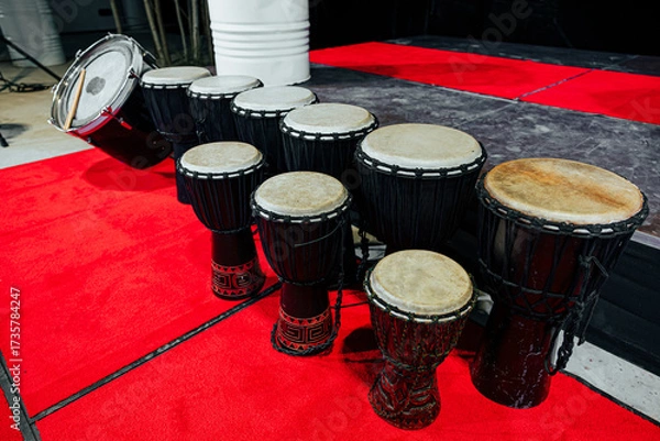 Fototapeta A line of traditional African djembes and a bass drum arranged on a red carpet stage, showcasing handcrafted design and cultural percussion instruments