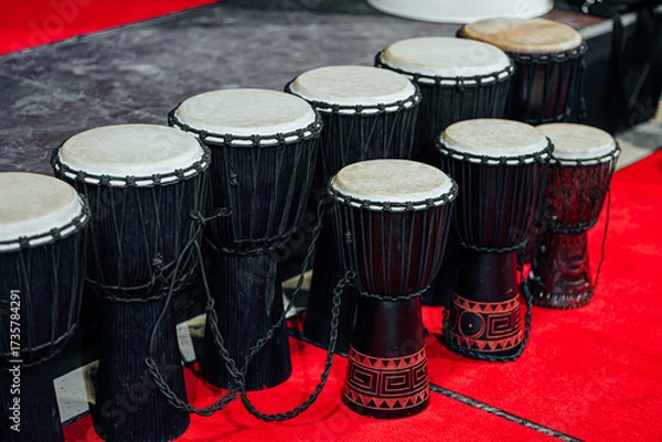 Fototapeta A line of traditional African djembes and a bass drum arranged on a red carpet stage, showcasing handcrafted design and cultural percussion instruments