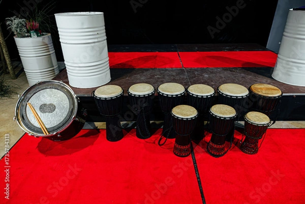 Fototapeta A line of traditional African djembes and a bass drum arranged on a red carpet stage, showcasing handcrafted design and cultural percussion instruments