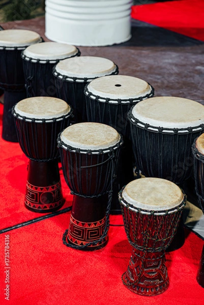 Fototapeta A line of traditional African djembes and a bass drum arranged on a red carpet stage, showcasing handcrafted design and cultural percussion instruments