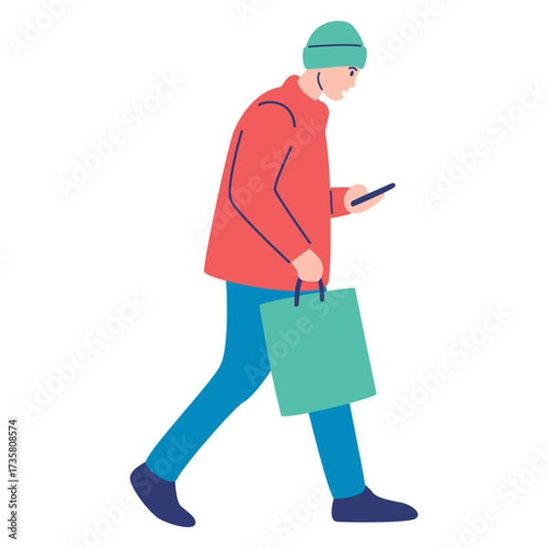 Fototapeta A young man in warm winter clothing walking with a shopping bag while looking down and using his smartphone, representing a modern urban lifestyle