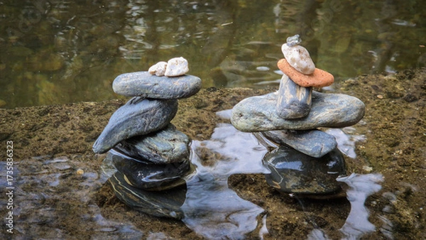 Fototapeta Stacks of balanced river pebbles on the water’s edge symbolize harmony, peace, and focus. Reflective surface and smooth stones evoke zen, mindfulness, and natural art.