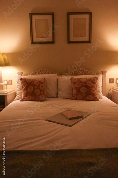 Fototapeta Cosy English bedroom at night with lit lamp, carved headboard, floral cushions and closed laptop with notebook on bed, concept of evening routine, home comfort and quiet retreat