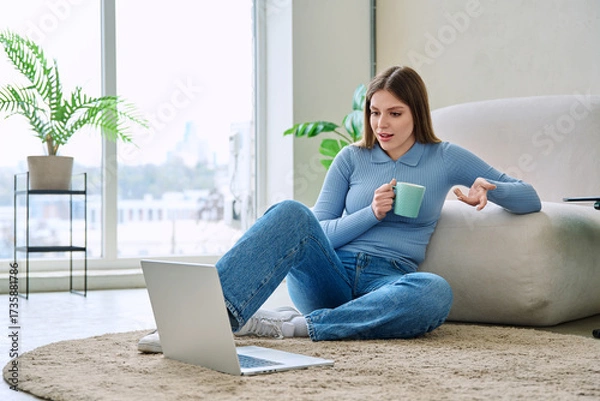 Fototapeta Young relaxed woman with cup using laptop, sitting on floor at home