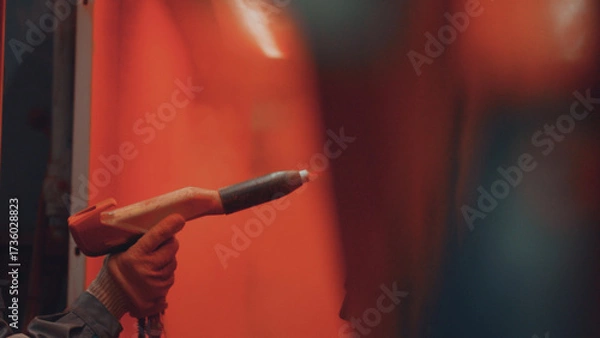 Fototapeta Worker hand applying a protective layer of powder coating to a metal part using a spray gun in a manufacturing workshop, showing process for surface finishing and durability