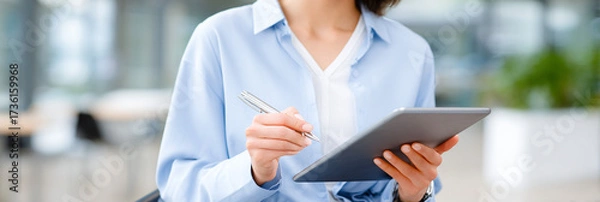 Fototapeta Professional woman in blue shirt holding tablet and pen, engaged in work, with modern office background showcasing a collaborative and productive environment