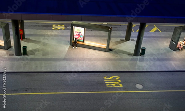 Fototapeta Urban bus station at night with illuminated shelters and a lone passenger waiting for transportation