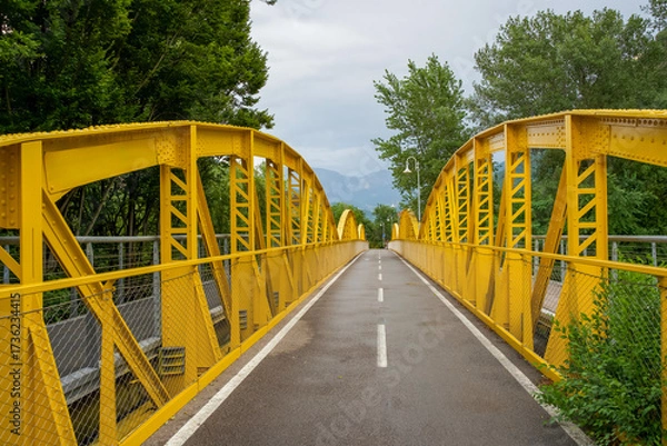 Fototapeta Yellow metal bridge spans over a road surrounded by lush greenery and mountains in the background with copy space