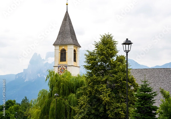 Fototapeta Historic church tower with clock surrounded by lush greenery and mountains in the background showcasing natural beauty