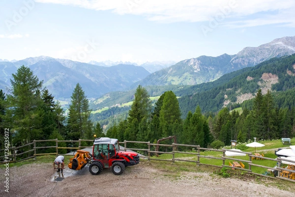 Fototapeta Red tractor parked on mountain landscape with worker preparing equipment in scenic alpine environment