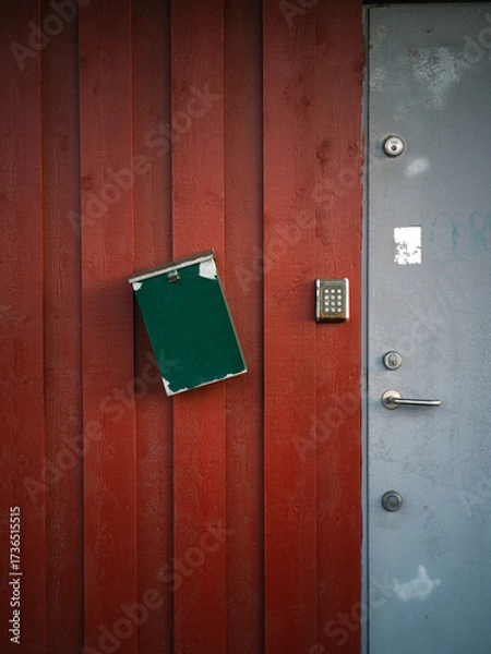 Obraz Red wall with grey steel door, key code and green mailbox hanging crooked 
