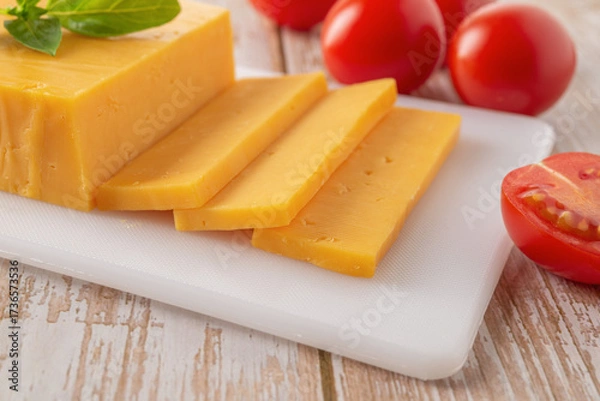 Fototapeta Sheddar cheese slices close-up. Slaced orange cheese on a white cutting board and few red tomatoes over wooden tabletop. Dairy product, calcium.