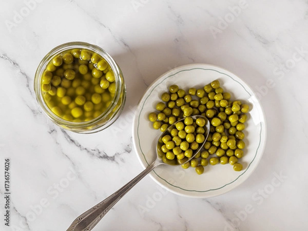 Fototapeta Canned green peas on a plate over marble countertop. Marinated organic peas on a white dish and spoon near glass jar. Vegetable protein, dietary fiber, salad ingredient.