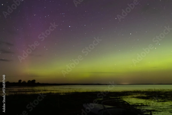 Fototapeta Bright green and red northern lights dance over a calm lake, casting vivid reflections on the water under a star-filled sky during a clear night.