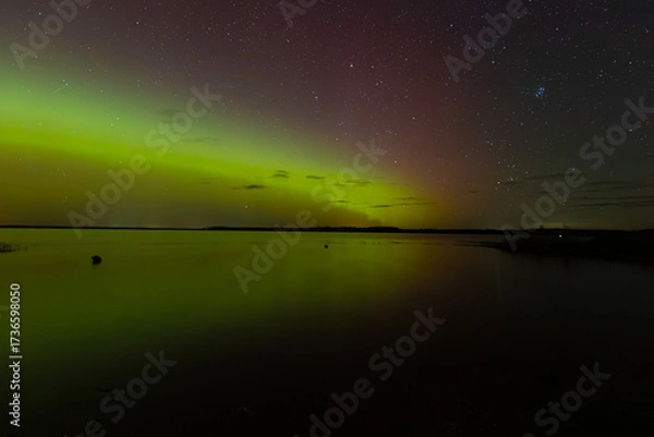 Fototapeta Bright green and red northern lights dance over a calm lake, casting vivid reflections on the water under a star-filled sky during a clear night.