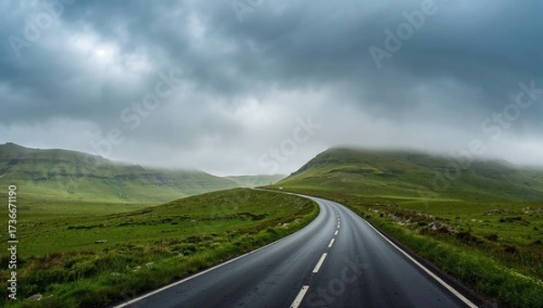 Fototapeta Long winding road cutting through lush green countryside under a moody cloudy sky
