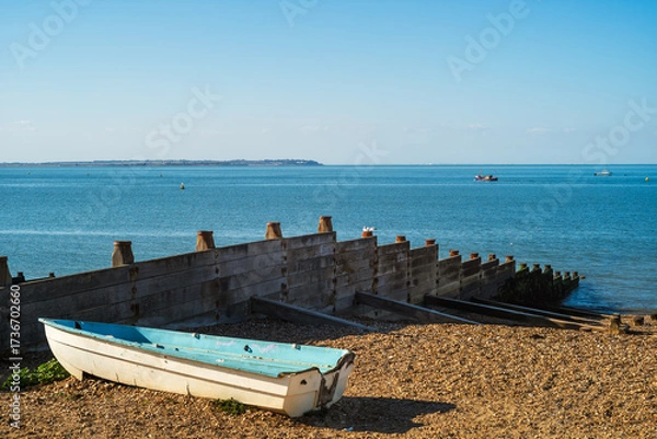 Fototapeta View across to the Isle of Sheppey from Whitstable beach. A wooden groyne and small blue boat are on the pebble beach.